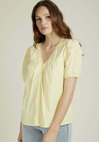 Blouse jaune avec des détails en broderie délicate sur les manches bouffantes, un décolleté en V et une finition en dentelle, assortie à un jean en denim clair.