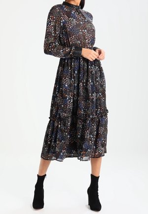 Vrouw draagt een lang zwart jurkje tot over de knie met kleurrijke bloemmotieven, een zwarte kraag en manchetten, gecombineerd met zwarte enkellaarsjes.