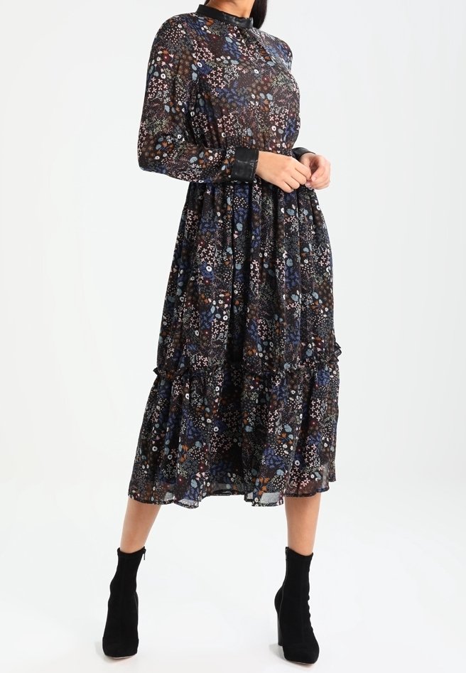 Femme portant une robe noire à manches longues, mi-longue jusqu'aux genoux, ornée de motifs floraux colorés, avec un col et des poignets noirs, associée à des bottines noires.