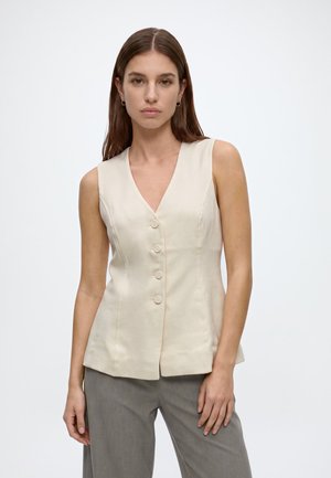 Frau mit glatten braunen Haaren trägt eine ärmellose beige Weste mit Knöpfen und graue Hose, steht vor einem schlichten weißen Hintergrund.