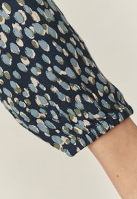 Tifon închis navy cu o suprafață texturată prezintă un model de pete albastre, verzi și roz. Mâneca are un detaliu cu o manșetă gathered.