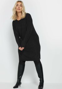 Vestito lungo in maglia nera con texture a costine e spacchi laterali; maniche lunghe e scollatura a V; abbinato a stivali neri in pelle al ginocchio.