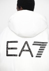 Balta mīksta jaka ar lielu melnu izšūtu "EA7" logotipu aizmugurē. Iekļauts kapuci un gluda, spīdīga tekstūra.