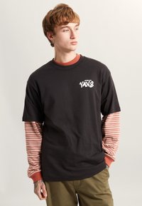 Zwarte oversized t-shirt met witte "VANS" logo, gelaagd over een roodgestreepte lange mouw, gecombineerd met khaki broek.