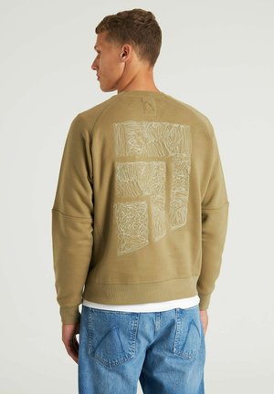 Sweatshirt vert olive avec un graphique texturé blanc dans le dos, présentant des manches longues et des poignets côtelés. Porté par-dessus une chemise blanche.