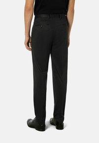 Homme portant un pantalon habillé gris foncé, une ceinture noire, une chemise noire et des chaussures habillées noires, vu de dos contre un fond clair.