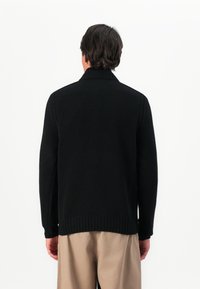 Homem com as costas viradas, vestindo um suéter de malha canelada preto e calças bege, contra um fundo branco simples.