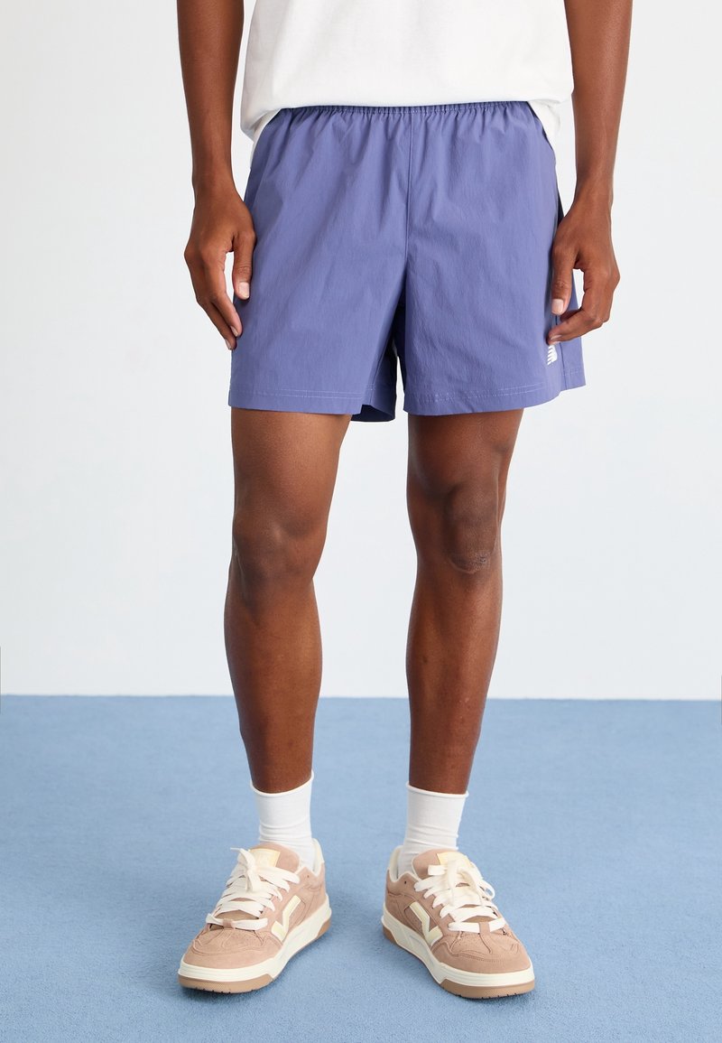 Homme portant un short de sport violet, des chaussettes blanches et des baskets beiges, debout sur un sol bleu devant un fond blanc.