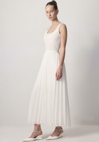 White pleated maxi dress with a fitted bodice, wide straps, and a smooth texture. Paired with white slingback shoes featuring a pointed toe.