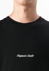 Close-up van een nek en bovenste borst die een zwart t-shirt draagt, met witte cursieve tekst die "Originals Studio" op de voorkant leest.