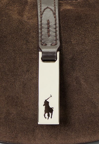 Braunes Wildledermaterial mit einem silbernen Metalletikett; das Etikett zeigt ein ausgeschnittenes Logo eines Pferdes und Reiters, das mit einem dunklen Lederband befestigt ist.