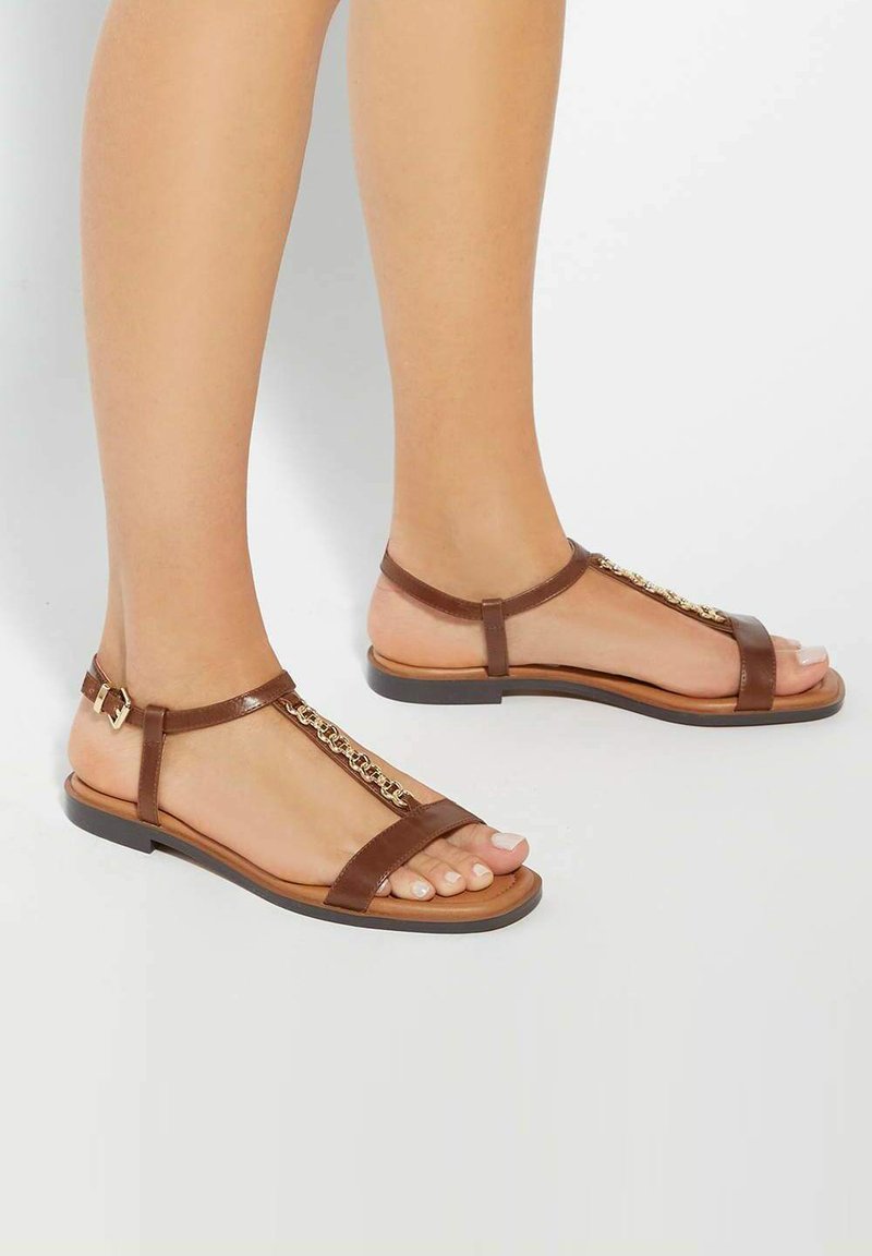 Brown leather sandals with a thin strap design, featuring a chain accent along the center and a buckle closure at the ankle. Flat sole.