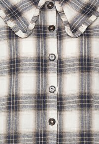 Camisa de flanela quadrada com padrões em azul-marinho, bege e creme; possui colarinho e seis botões escuros na frente. Textura suave.