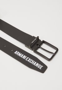 Cintura nera con finitura opaca, dotata di una fibbia metallica rettangolare e testo bianco "ARMANI EXCHANGE" sulla cintura.