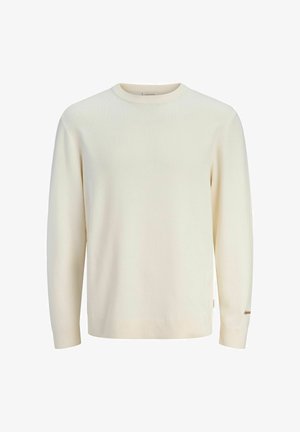 Teismeline poiss, kes kannab rohelist jopet, valget kampsunit, lahtisi siniseid teksapükse, beežide kingi ja musta nokamütsi, seisab valge ühevärvilise tausta ees.