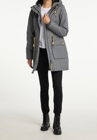 Šedá parka s texturovaným povrchem, nastavitelnými žlutými šňůrkami, dvěma předními kapsami a kapucí. Nošena přes černé kalhoty a boty.