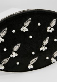 Pochette ovale noire avec motifs floraux en grappes de perles argentées et une fermeture éclair argentée le long du bord supérieur.