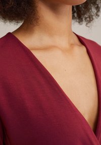 Burgundy huppari, joka on valmistettu sileästä kankaasta, syvällä V-pääntie ja lyhyet hihat, korostaen pehmeää tekstuuria ja selkeitä linjoja.