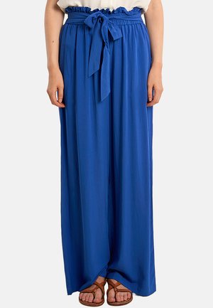 Femme portant un pantalon taille haute, jambes larges, bleu roi, avec une ceinture en tissu nouée en nœud papillon, associé à des sandales marron à lanières.