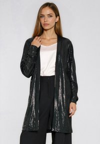 Czarny, długi kardigan z cekinami, o lekkim, otwartym fasonie, z długimi rękawami i gładką fakturą. Noszony na jasnym topie.