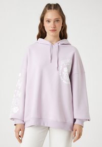 Jasnofioletowa hoodie z przednią kieszenią, oversize'owym krojem, kapturem ze sznurkiem oraz białymi grafikami kwiatowymi na rękawie i klatce piersiowej.