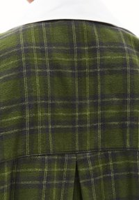 Tessuto a quadri verdi e neri con dettagli di cucitura. Presenta un colletto bianco e una piega sul retro per un tocco di stile e movimento.