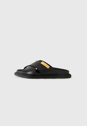 Black slide sandal with wide crossed straps and a small rectangular yellow label on one strap, displayed against a plain white background.