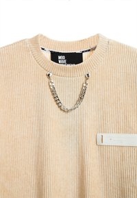 Sudadera de pana beige con textura acanalada, presenta un detalle de cadena plateada y una etiqueta rectangular con texto en relieve en el dobladillo.