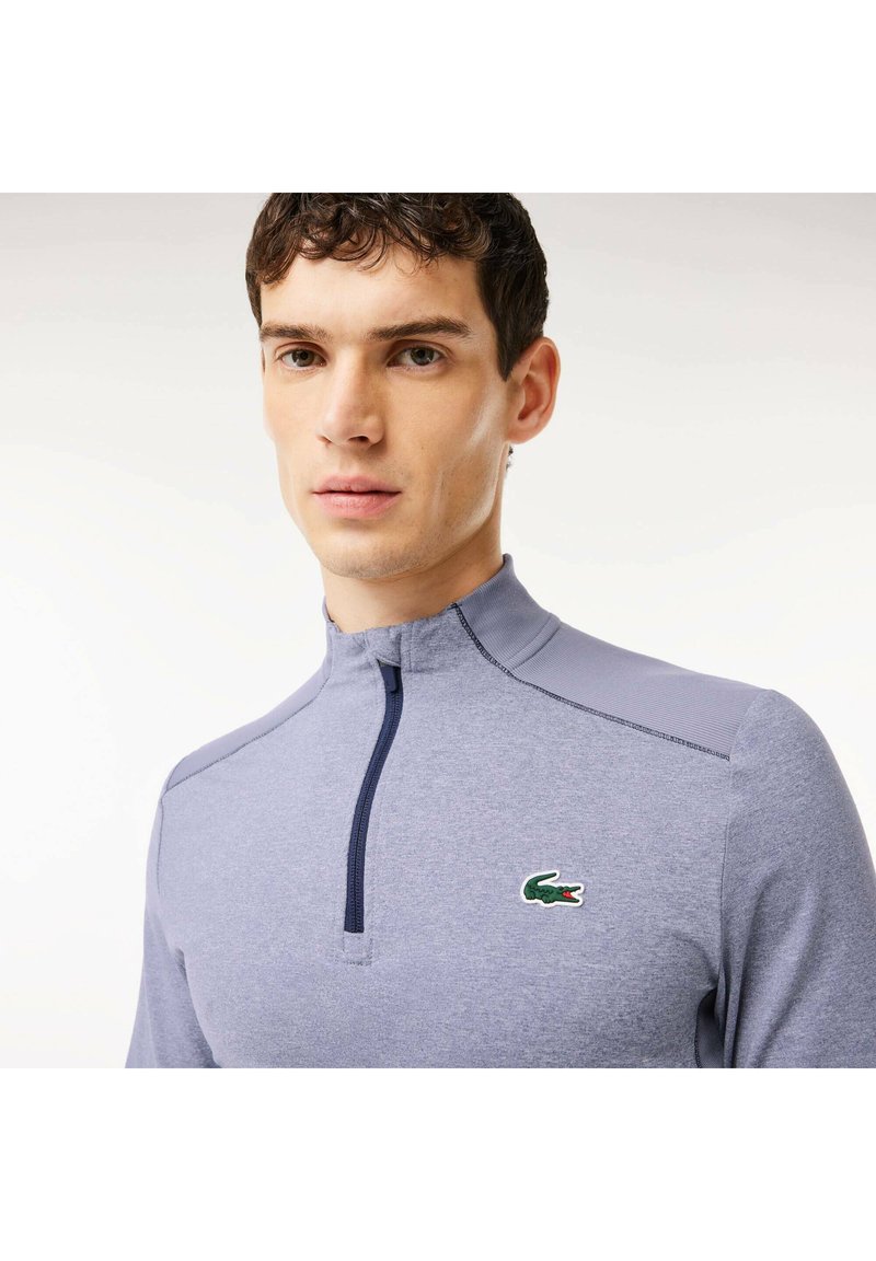 Herren Pullover in Grau mit Viertelreißverschluss, strukturiertem Stoff, kontrastierenden Nähten und einem kleinen grünen Krokodil-Logo auf der Brust.