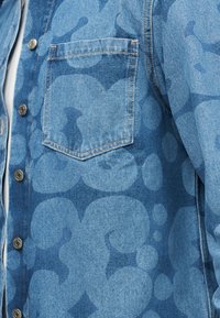 Μπλε πουκάμισο από τζιν με κούμπωμα με κουμπιά, τσέπη στο στήθος και μεγάλο αφηρημένο κυκλικό μοτίβο σε διάφορες αποχρώσεις του μπλε.