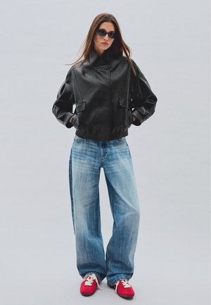 Kvinde iført sort læderjakke, løsthængende falmede blå jeans, røde sneakers og sorte solbriller, stående med hænderne i lommerne.