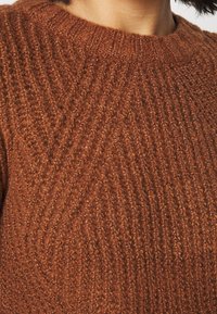 Gros plan d'une personne portant un pull marron texturé en maille côtelée avec un col rond et des cheveux bouclés foncés visibles.