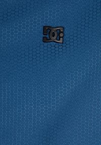 Blå strukturerad tyg med ett hexagonalt mönster, med en svart DC-logotyp som är tydligt visad i mitten.