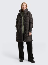 Fekete steppelt kabát magas gallérral, cipzáras és gombos záródással, steppelt mintával és oldalzsebekkel, zöld pulóver és fekete nadrág fölött viselve.