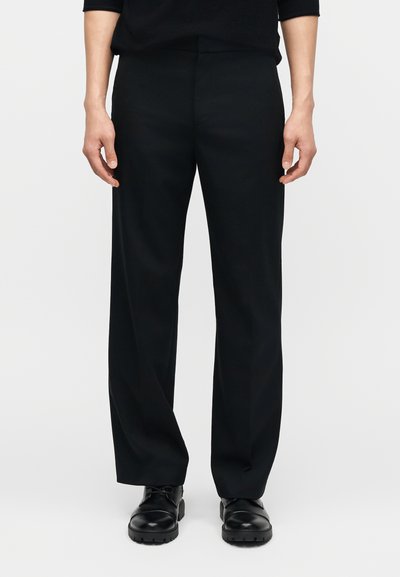 Άτομο που φοράει μαύρο παντελόνι ίσιας γραμμής και μαύρα δερμάτινα παπούτσια με κορδόνια, όρθιο μπροστά σε απλό ανοιχτό φόντο.