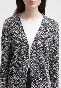 Dzianinowy kardigan w czarno-białe geometryczne wzory, o otwartym przodzie i długich rękawach. Miękka tekstura i uniwersalny styl.