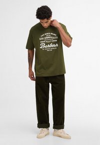 T-shirt en coton vert olive avec texte blanc imprimé. Associé à un pantalon en velours côtelé vert foncé et des baskets de couleur claire. Coupe décontractée.