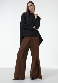 Top de manga larga texturado en negro combinado con pantalones de pierna ancha marrones. Los pantalones tienen un pliegue frontal marcado y se combinan con zapatos negros.