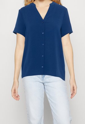 Mujer con blusa azul marino de manga corta y botones, con escote en V, combinada con vaqueros azul claro, sobre un fondo liso.
