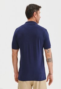 Granatowa koszulka polo wykonana z miękkiego materiału, z krótkimi rękawami i klasycznym kołnierzykiem, widoczna z tyłu, z widocznymi detalami tatuażu.
