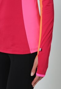 Długi rękaw sportowego topu o jasnoczerwonym i różowym materiale, z kontrastowym pomarańczowym bocznym panelem, prążkowaną fakturą oraz logiem na rękawie.
