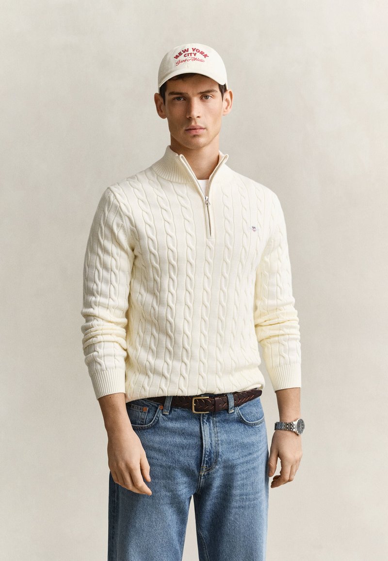 Jeune homme portant un pull en maille torsadée blanc avec fermeture éclair, jean bleu, montre argentée, ceinture marron et casquette blanche avec logo New York City rouge.