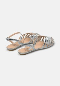 Silberne Sandalen mit Riemchen, die ein offenes Design, eine flache Sohle und einen verstellbaren Knöchelriemen aufweisen. Glatte, metallische Oberfläche mit minimalistischen Details.