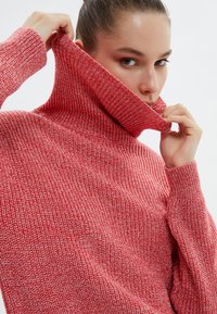 Pull en maille rouge texturé, à col roulé haut, avec des poignets côtelés et une coupe décontractée. La vue en gros plan met l'accent sur les détails du matériau.