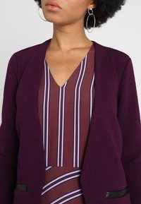 Lila texturerad kavaj med en v-ringad randig blus som har mörkröda och lavendelfärgade vertikala ränder. Svarta detaljer på fickorna.