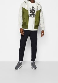 Giacca a vento bianca e verde oliva con cappuccio, abbinata a una t-shirt bianca con un design grafico nero, pantaloni scuri e sneakers grigie.