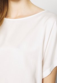 Luźna bluzka z krótkim rękawem, wykonana z miękkiego, lekkiego białego materiału, z szerokim, okrągłym dekoltem i gładką, niestrukturowaną fakturą.