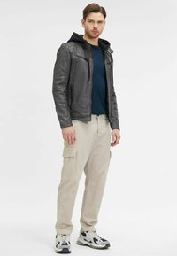 Giubbotto di pelle grigio scuro con cappuccio nero, abbinato a pantaloni cargo beige chiaro e scarpe sportive con un motivo di colori misti.