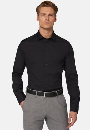 Schwarzes langärmliges Hemd mit spitzem Kragen und Knopfverschluss, kombiniert mit grauen gemusterten Stoffhosen und einem schwarzen Gürtel.