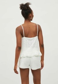 Pantaloni corti bianchi in cotone e una maglietta senza maniche con dettagli ad occhio, caratterizzata da un orlo a balze. Semplice, leggera, con sottili spalline.
